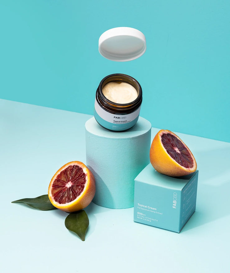 FABCBD topical cream jar with open lid floating above a pedestal beside its teal box and halved blood oranges