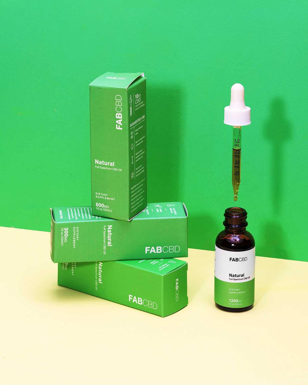Three green FABCBD Natural Full Spectrum CBD Oil retail boxes in varying strengths (300mg, 600mg, and 1200mg) stacked and arranged on a cream surface next to an amber dropper bottle of FABCBD Natural Full Spectrum CBD Oil (1200mg) with the dropper pulled out and held above it, against a two-tone green and cream background.